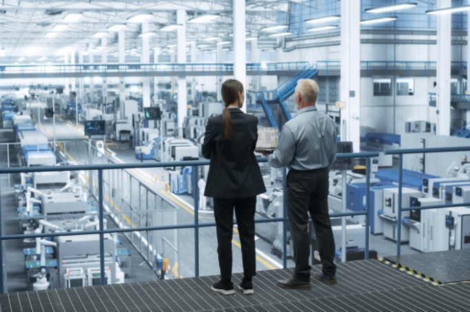 Two people stand on a balcony overlooking a large, modern factory floor filled with industrial machinery. The atmosphere is industrious and focused.